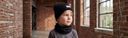 Chłopiec w komplecie „Oskar” – czapka beanie i komin – w kolorze Czarny stoi w ceglanej hali przy dużych oknach.