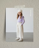 Młoda dziewczyna z długimi kręconymi włosami ma na sobie białą czapkę, jasnofioletową bluzkę z długim rękawem i falbanami, białe spodnie z szerokimi nogawkami i białe trampki. Stoi w pomieszczeniu na prostym tle, patrząc lekko w bok.