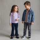 Dwoje małych dzieci stoi obok siebie, uśmiechając się do siebie. Dziewczynka ma długie kręcone włosy, ubrana jest w lawendowy top i granatowe spodnie. Chłopiec ma na sobie koszulę w paski, dżinsową kurtkę, szare dżinsy i czarne trampki. Tło neutralne.