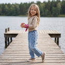 Uśmiechnięta młoda dziewczyna z długimi włosami spaceruje po drewnianej przystani nad jeziorem, ubrana w beżowy sweter, niebieskie dżinsy, buty w kropki i różowy szalik na nadgarstku. W tle widoczne są drzewa.