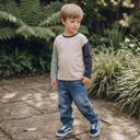 Młody chłopak o jasnych włosach stoi na kamiennej ścieżce w ogrodzie, ubrany w kolorową koszulę z długim rękawem, niebieskie dżinsy i granatowe trampki, patrząc w bok otoczony bujną zielenią roślin.