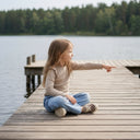 Młoda dziewczyna z długimi włosami, ubrana w beżowy top i niebieskie dżinsy, siedzi ze skrzyżowanymi nogami na drewnianym pomoście nad jeziorem, wskazując w kierunku wody. W tle widoczne są drzewa i inna część doku.