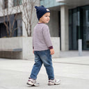 Małe dziecko ubrane w koszulę w paski, dżinsy, trampki i granatową czapkę z uszami niedźwiedzia spaceruje na zewnątrz w pobliżu nowoczesnych budynków i betonowych donic.