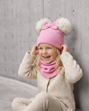 Dziewczynka w komplecie „Pom Pom” w kolorze Baby pink śmieje się, trzymając czapkę obiema dłońmi, i siedzi przy betonowej ścianie.