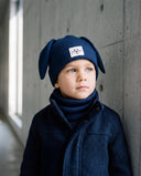 Chłopiec w komplecie „Frost” w kolorze Jeans stoi przy betonowej ścianie, zbliżenie na czapkę i komin.