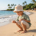 Małe dziecko w czapce typu bucket hat „PanPan 35 Kapelusz muślinowy chłopięcy” oraz w lekkim ubranku z muślinu bawełnianego kuca na słonecznej plaży, przyglądając się muszli, a w tle widać delikatne fale i palmy.