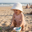 Małe dziecko w jasnoróżowym stroju uśmiecha się i bawi się na piaszczystej plaży, mając na głowie muślinową czapeczkę PanPan 35 z kokardką; w tle widać wiaderko, łopatkę, ocean i ludzi.