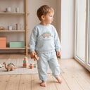 Małe dziecko ubrane w zestaw chłopięcy PanPan 35 „Dres z dinozaurem” – bluzę i spodnie z bawełny – stoi przy oknie, trzymając w ręku drewnianą zabawkę. W przytulnym pokoju widać drewniane klocki, figurkę dinozaura oraz półki z przedmiotami dekoracyjnymi – to idealne otoczenie dla odzieży do przedszkola.