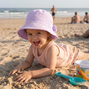 Uśmiechnięte maluch w różowym ubranku i czapeczce muślinowej PanPan 35 z kokardką leży na piaszczystej plaży, bawiąc się piaskiem obok kolorowego wiaderka i łopatki. W tle widać fale oceanu i kilka osób.