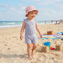 Uśmiechnięta maluch w muślinowym kapeluszu dziewczęcym „PanPan 35” z kokardką i cyrkoniami oraz w pasiastym kombinezonie spaceruje boso po piaszczystej plaży w pobliżu zamków z piasku, z zabawkami, koszem piknikowym i kolorowym kocem. W tle widać ocean i niebo.