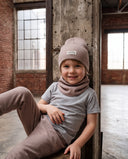 Uśmiechnięty chłopiec w zestawie czapka beanie i komin siedzi oparty o betonowy filar w hali.