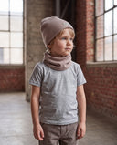 Chłopiec w zestawie czapka beanie i komin stoi w hali przy oknach i patrzy w bok.