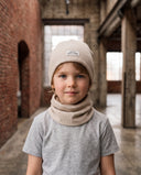 Chłopiec w zestawie czapka beanie i komin stoi w hali i patrzy w obiektyw, w tle ceglany mur.