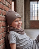 Śmiejący się chłopiec w zestawie czapka beanie i komin siedzi oparty o ceglany mur w hali.