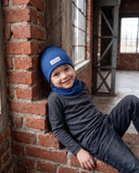 Uśmiechnięty chłopiec w zestawie czapka beanie i komin siedzi oparty o ceglany mur.