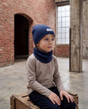 Chłopiec w zestawie czapka beanie i komin siedzi na drewnianej skrzyni w hali i patrzy w bok.