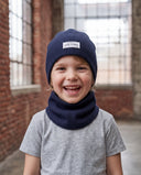 Zbliżenie na śmiejącego się chłopca w czapce beanie i kominie, w tle ceglany mur i okno.