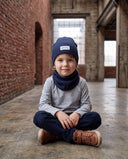 Uśmiechnięty chłopiec w zestawie czapka beanie i komin siedzi po turecku na podłodze hali.