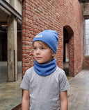 Chłopiec w zestawie czapka beanie i komin stoi w hali przy ceglanym murze i patrzy w bok.