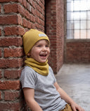 Śmiejący się chłopiec w zestawie czapka beanie i komin siedzi oparty o ceglany mur.