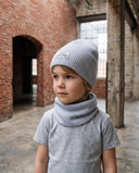 Chłopiec w zestawie czapka beanie i komin stoi w hali i patrzy w bok, w tle ceglany łuk.