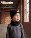 Chłopiec w zestawie czapka beanie i komin stoi przy dużych oknach ceglanej hali i patrzy w bok.