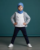 Młody chłopak pozuje pewnie w PanPan 35 Zestaw czapka beanie i komin dla chłopca - bawełna, prążek, wiosna z jasnoszarą kurtką, białą koszulą, ciemnoniebieskimi spodniami i białymi trampkami na jednolitym turkusowym tle.