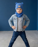 Młody chłopak nosi PanPan 35 Wiosenny komplet miś dla chłopca - czapka z uszkami i komin, wraz z dżinsową kurtką i dżinsami, pozując pewnie w minimalistycznym pokoju o niebieskich ścianach z betonową podłogą.