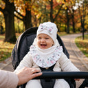 Uśmiechnięte dziecko w wózku ma na sobie zestaw PanPan 35 Komplet dla dziewczynki „Rosalia” — białą czapkę z prążkowanej bawełny oraz chustę z delikatnymi kwiatowymi wzorami — podczas spaceru w parku w słoneczny jesienny dzień. Ręka osoby dorosłej spoczywa na rączce wózka.