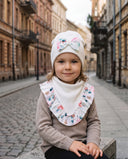 Dziewczynka w czapce z kokardą i chuście w kwiatowy print siedzi na kamiennym murku przy brukowanej ulicy.