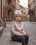 Dziewczynka w czapce z kokardą i chuście w kwiatowy print siedzi na kamiennym murku przy brukowanej ulicy.