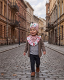Dziewczynka w czapce z kokardą i chuście w kwiatowy print idzie uśmiechnięta brukowaną ulicą.
