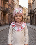 Dziewczynka w czapce z kokardą i chuście w kwiatowy print stoi na brukowanej ulicy, patrząc w obiektyw.
