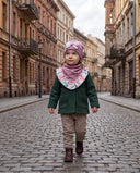 Dziewczynka w czapce z kokardą i chuście w kwiatowy print idzie brukowaną ulicą wzdłuż kamienic.