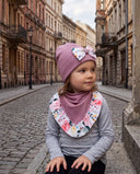 Dziewczynka w czapce z kokardą i chuście w kwiatowy print siedzi na kamiennym progu przy brukowanej ulicy.