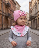 Dziewczynka w czapce z kokardą i chuście w kwiatowy print stoi na brukowanej ulicy, patrząc w bok.