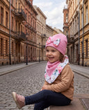 Dziewczynka w czapce z kokardą i chuście w kwiatowy print śmieje się, siedząc na brukowanej ulicy.