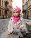 Dziewczynka w czapce z kokardą i chuście w kwiatowy print siedzi na kamiennym murku przy brukowanej ulicy.