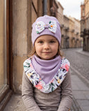 Dziewczynka w czapce z kokardą i chuście w kwiatowy print, zbliżenie na twarz przy ścianie budynku na miejskiej ulicy.