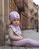 Dziewczynka w czapce z kokardą i chuście w kwiatowy print siedzi na chodniku przy kamiennym słupie, w tle kamienice.