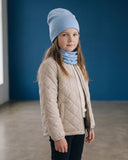 Młoda dziewczyna z długimi brązowymi włosami stoi w pomieszczeniu, ubrana w PanPan 35 Wiosenny komplet dla dziewczynki - czapka z perełkami i komin oraz beżową pikowaną kurtkę, na tle niebieskiej ściany i dużego okna, patrząc neutralnie w kamerę.