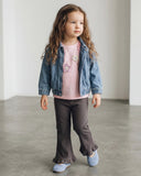 Młoda dziewczyna z długimi brązowymi włosami stoi w pomieszczeniu na szarej podłodze, ubrana w jeansową kurtkę dziecięcą unisex PanPan 35 - wiosenna, przejściowa na różową koszulę w kwiaty, ciemne marszczone spodnie i jasnoniebieskie buty, patrząc w bok.