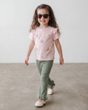 Małe dziecko z długimi kręconymi włosami, w okularach przeciwsłonecznych, zielonych spodniach z marszczonymi obszyciami, kremowych butach i koszulce PanPan 35 Bawełniany T-shirt dla dziewczynki z kwiatkami 3D i falbanką na rękawkach idzie pewnie po betonowej podłodze.