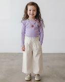 Młoda dziewczyna z długimi kręconymi włosami uśmiecha się w PanPan 35 bluzka dziewczęca z falbanką z wiskozy i elastanu (długi rękaw), kremowe spodnie z szerokimi nogawkami, wzorzyste buty i kolorowy naszyjnik z koralików na jednolitym tle.