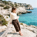Młoda dziewczyna stoi boso na skalistym wybrzeżu, opierając się o gałąź wyrzuconą przez morze. Ma na sobie bluzkę dziewczęcą PanPan 35 z falbanką, wykonaną z wiskozy i elastanu, z długim rękawem w kolorze kremowym, w połączeniu z miękkimi brązowymi spodniami z dzianiny wiskozowej. W tle, pod czystym niebem, widać klify i turkusową wodę.