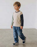 Chłopiec o jasnobrązowych włosach ma na sobie blok kolorystyczny PanPan 35 Bawełniana bluzka chłopięca z długim rękawem, niebieskie dżinsy i trampki w kratkę, stojąc na gładkiej, jasnej podłodze przy gładkiej białej ścianie.