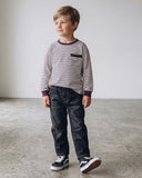 Młody chłopak stoi w pomieszczeniu na wypolerowanej betonowej podłodze, ubrany w PanPan 35 bawełniana bluzka chłopięca w paski z kieszonką i nadrukiem kota, ciemne dżinsy i czarno-białe trampki, patrząc w bok z lekkim uśmiechem.