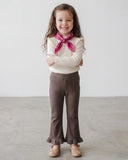 Młoda dziewczyna z długimi brązowymi włosami uśmiecha się, krzyżując ręce, ubrana w kremową bluzkę z długim rękawem, PanPan 35 Wiosenne leginsy dzwony z falbanką dla dziewczynki, brązowe buty w kwiaty i różowy szalik. Stoi na szarej podłodze przy białej ścianie.