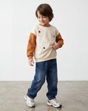 Małe dziecko z brązowymi włosami do ramion stoi na betonowej podłodze, uśmiechając się w bok. Ubrane jest w beżowy sweter z brązowymi rękawami, białe trampki i PanPan 35 jeansy chłopięce z gumką w pasie i imitacją rozporka.