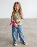 Młoda dziewczyna z długimi blond włosami stoi uśmiechnięta na betonowej podłodze. Ma na sobie beżową koszulę z długim rękawem, PanPan 35 jeansy dziewczęce z szeroką nogawką i haftowanymi wisienkami, buty w kratkę i różową apaszkę na nadgarstku.